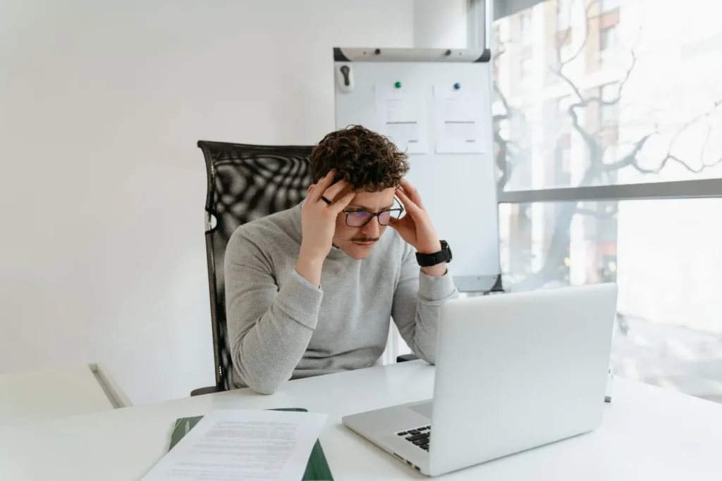 A tense man sits in front of a laptop with his hands on his temples
