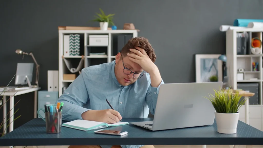 Mann schreibt mit der Hand an der Stirn neben einem Laptop in ein Notizbuch