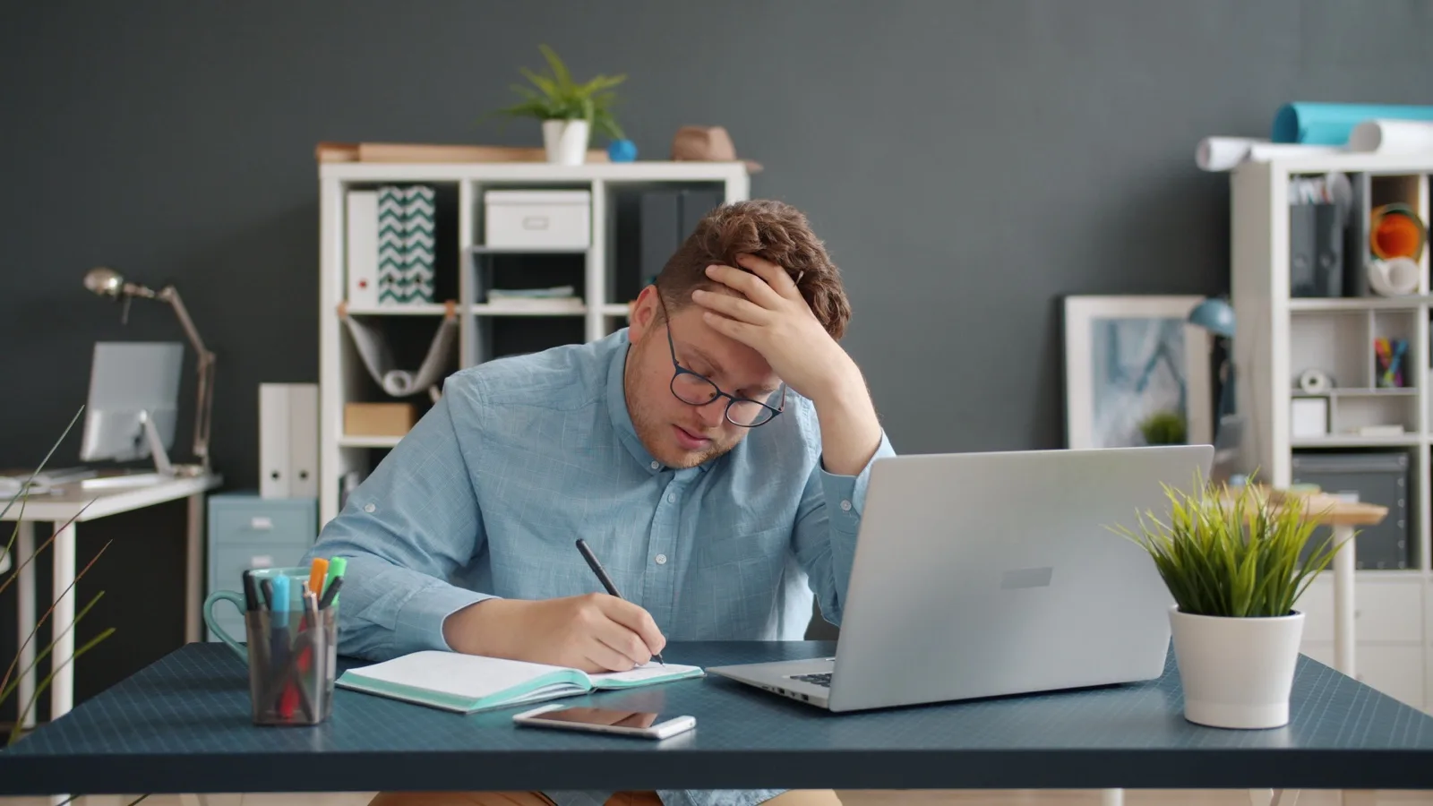 Mann schreibt mit der Hand an der Stirn neben einem Laptop in ein Notizbuch