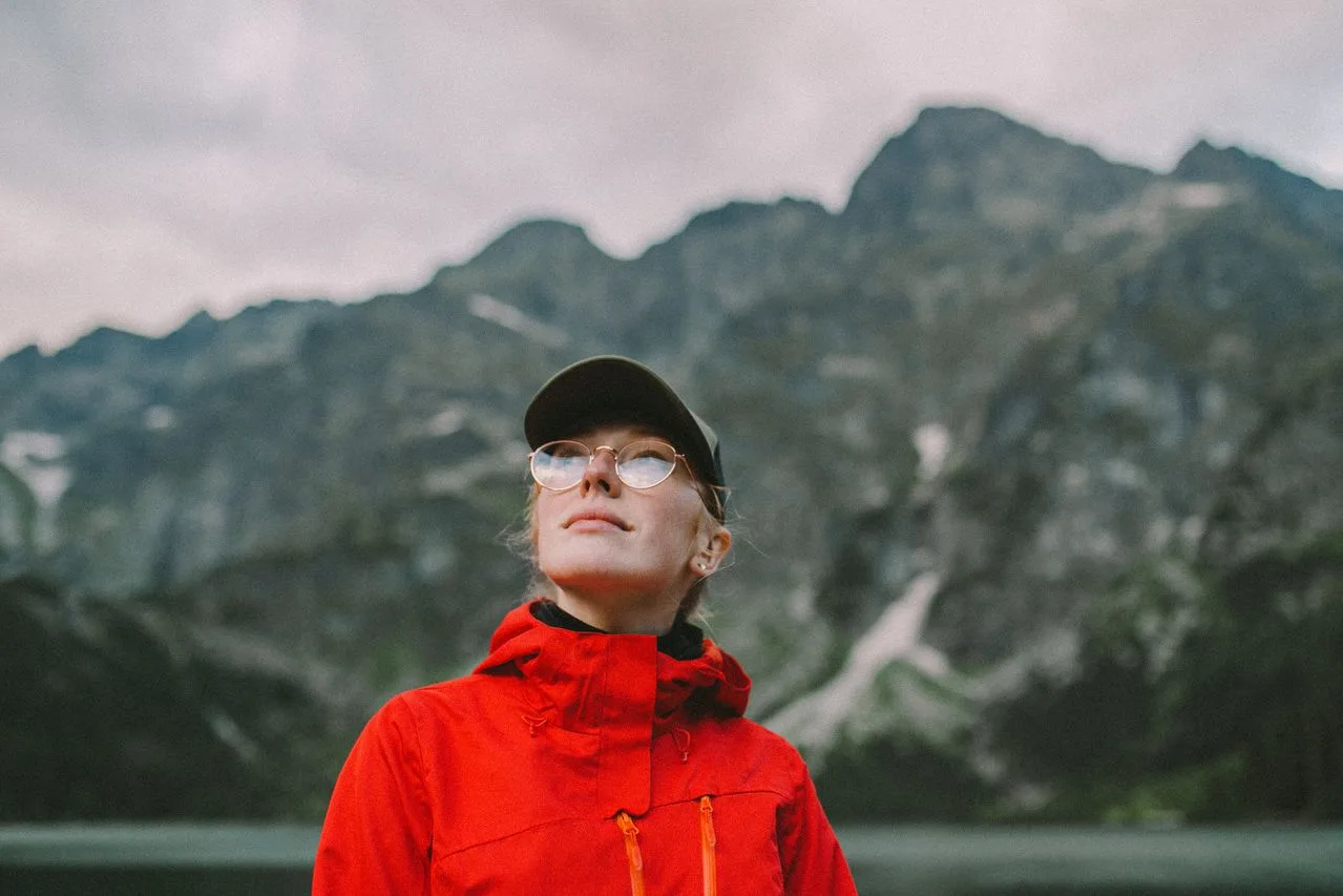 A woman in a red jacket and glasses is standing in front of a mountain landscape